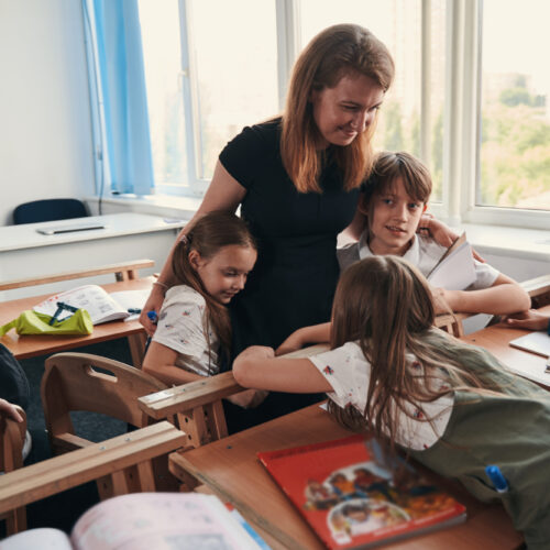 Smiling woman standing in a classroom and smiling while embracing schoolchildren by their shoulders